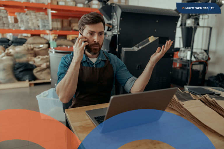 Commerciante preoccupato al telefono davanti al computer nel suo negozio, simbolo delle difficoltà economiche e dell’incertezza del mercato.